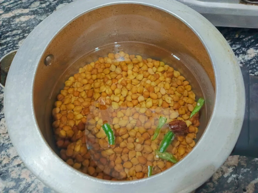 First of all, add kala chane (black chickpeas), 1 black cardamom (black ilaychi), and a small stick of cinnamon (daal chini) in the pressure cooker and add enough water. Now close the lid and cook for 6-7 whistles until the chickpeas become almost soft but still a little firm.